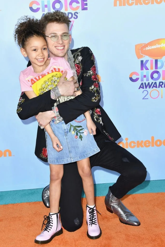 Person in floral jacket hugging a smiling child in a denim skirt on a red carpet at a Nickelodeon event