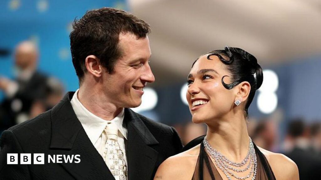 Dua Lipa and Callum Turner smile at each other - she wears a multi-stranded sparkling necklace and halterneck outfit, and has slicked back hair styled in curls around her face, while Turner wears a dark suit and cream tie and shirt