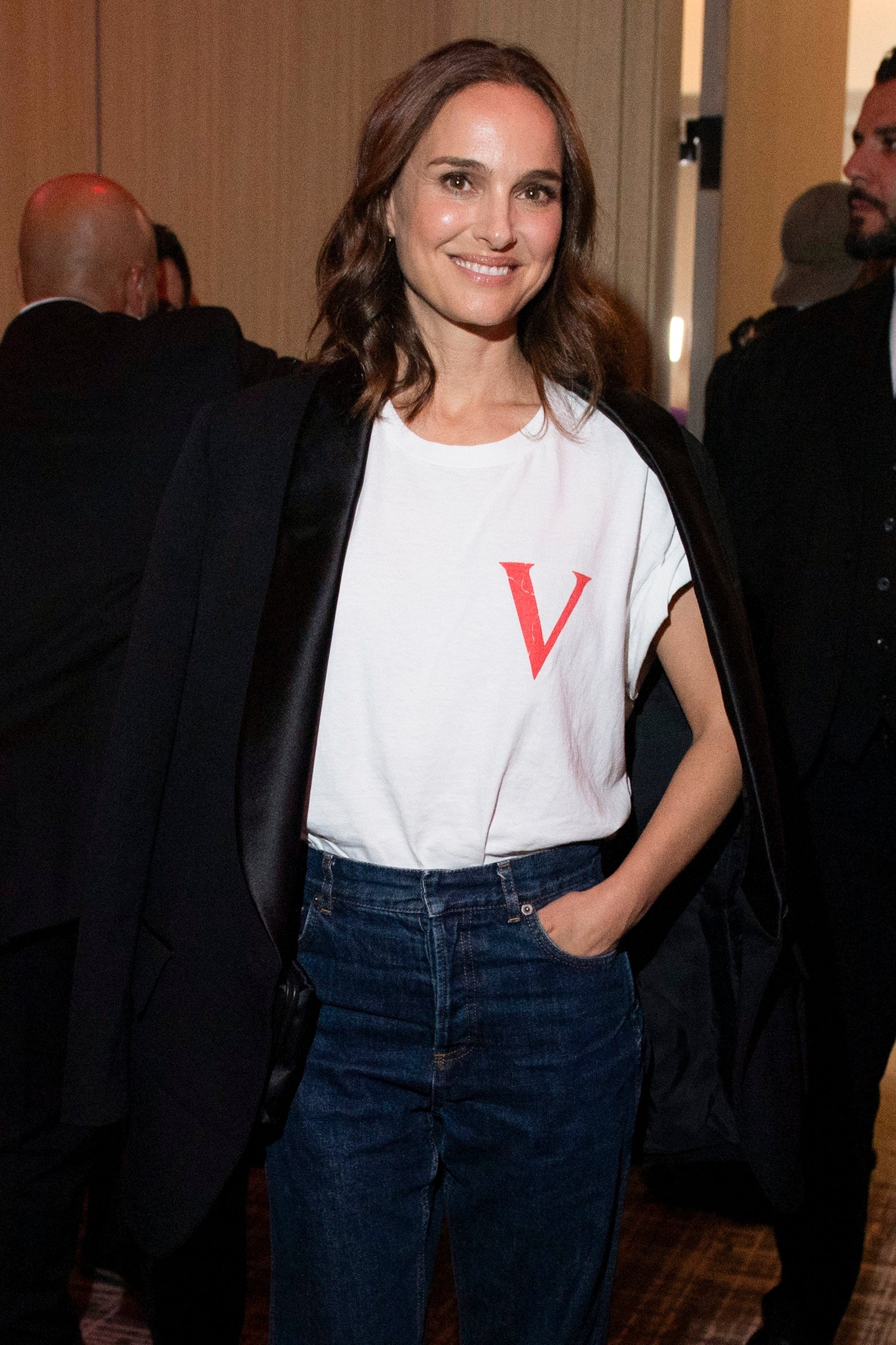 Person at an event wearing a casual outfit: a white T-shirt with a “V” and jeans, draping a blazer over their shoulders. Smiling at the camera