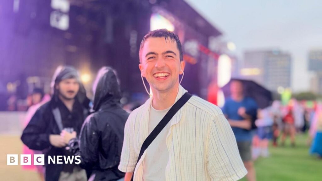 Alex Milsom is wearing a wet striped t-shirt and has wet hair. He is standing in a festival area in front of a stage with other people wearing rainproof jackets.