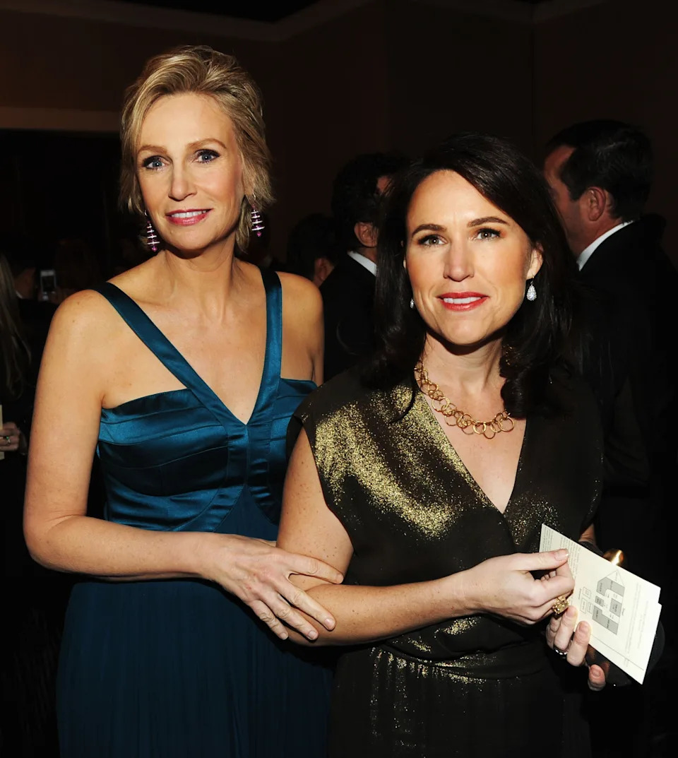 Two women at an event, one in a sleeveless gown and the other in a sparkly dress, standing closely and smiling at the camera