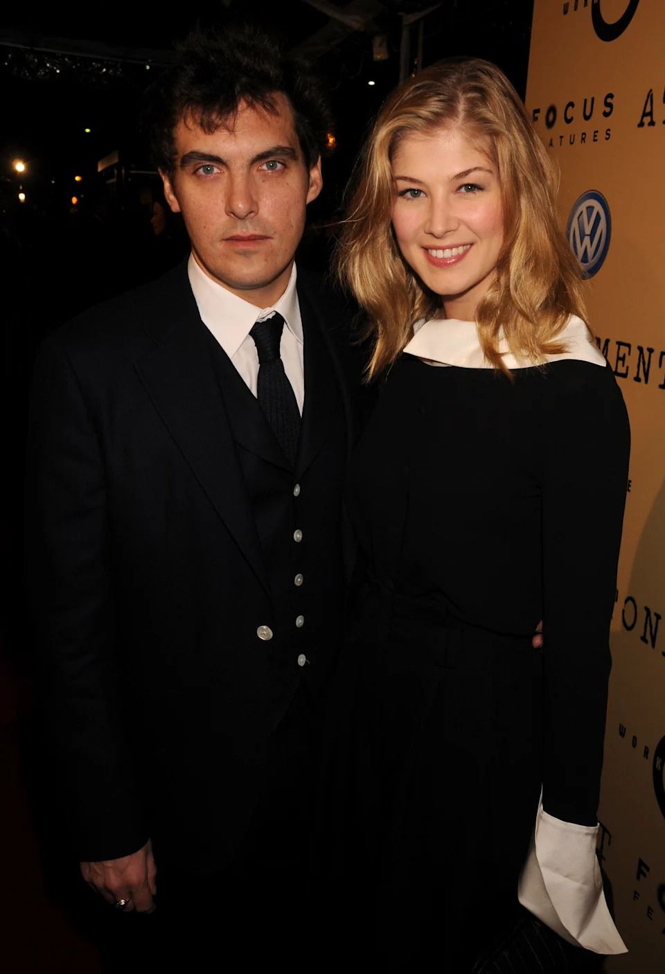 Two people on a red carpet. The woman wears a black dress with a white collar, and the man wears a dark suit with a tie