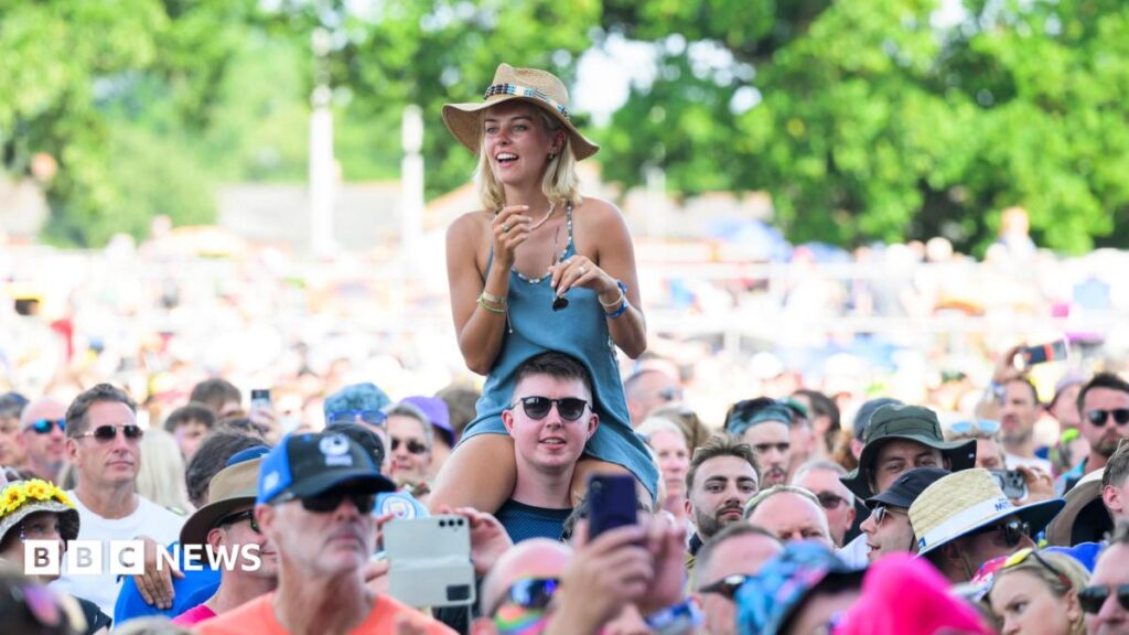 Your guide to the Isle of Wight Festival 2026 A music festival crowd is pictured head on. In the centre of the image a blonde woman wearing a blue dress is sat on a man's shoulders and towering over everybody else.