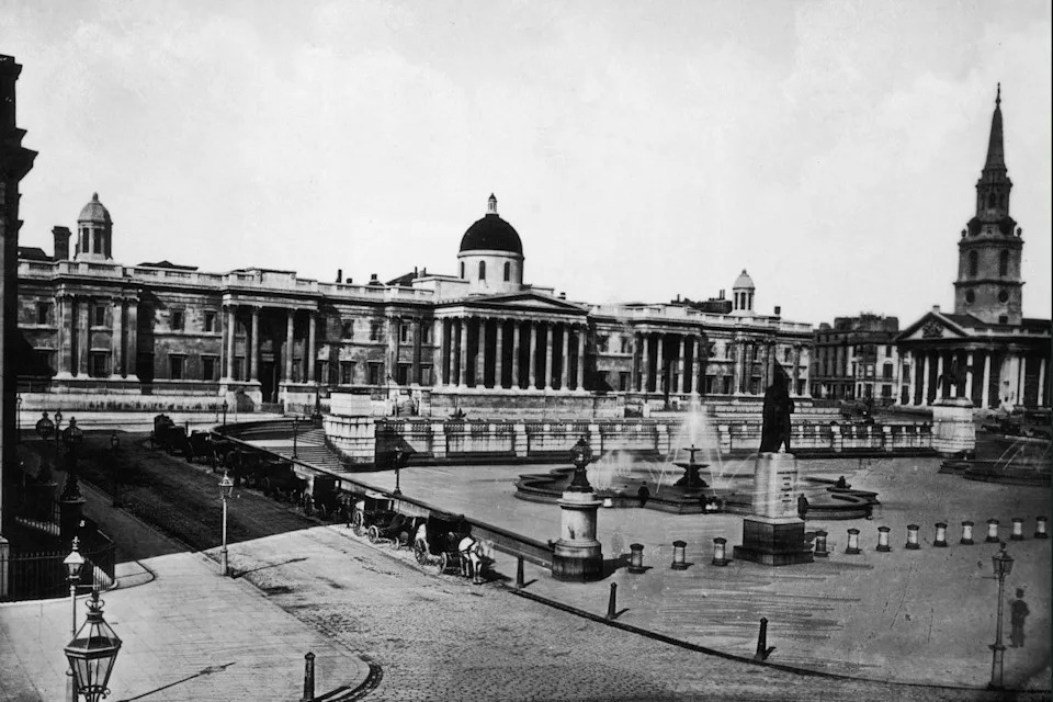 A black and white image from 1886 showing Trafalgar Square with the National Gallery along the back of the square beside St Martin-in-the-Fields. Horse and carts are lined up along one road