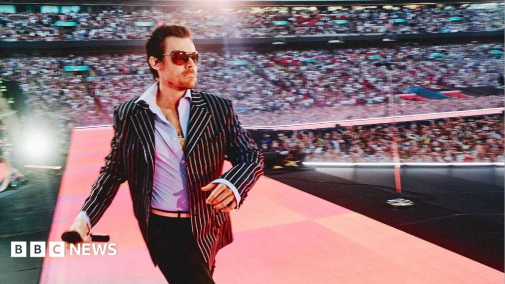 Harry Styles revisits X Factor as he kicks off Wembley residency Harry Styles strides onto the stage at Wembley Stadium, wearing a black and white pinstripe jacket and a wide-collared blue shirt. Spotlights illuminate the audience, and the singer is wearing aviator sunglasses.