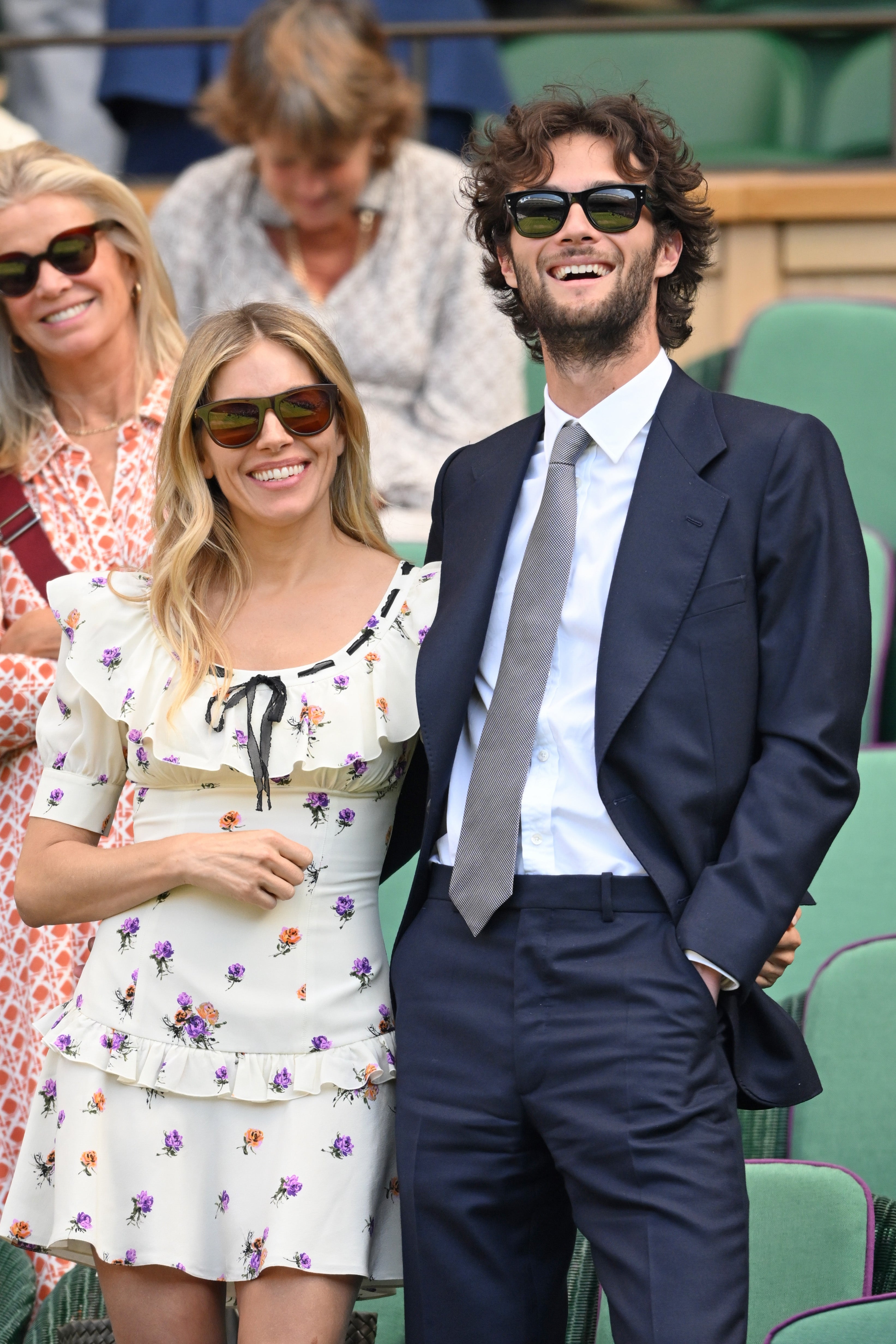 Two people smiling at an outdoor event; one in a floral dress, the other in a suit and tie, both wearing sunglasses