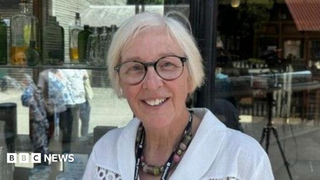 A woman with grey hair, glasses and a white blouse is sitting in front of a large window.