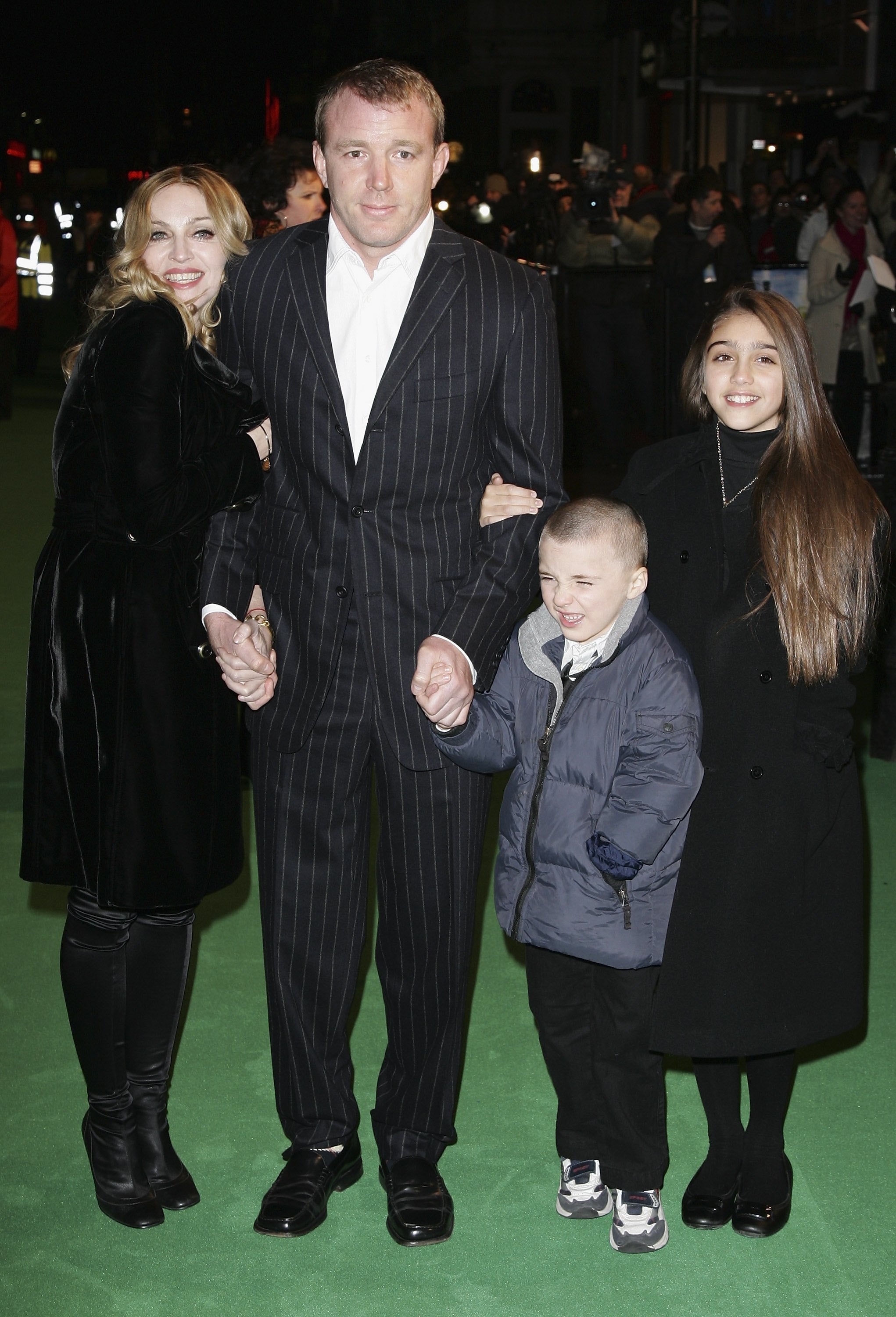 A family on a green carpet: two adults in formal clothing and two children in casual attire, posing together and smiling for cameras