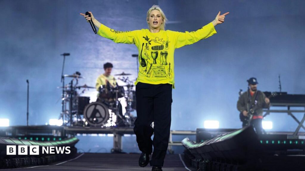 Download 2026: Linkin Park ignore the noise with triumphant set A woman wearing a yellow top and holding a microphone in her left hand is on stage, walking towards the camera. In the background behind her, both left and right, are band members playing.