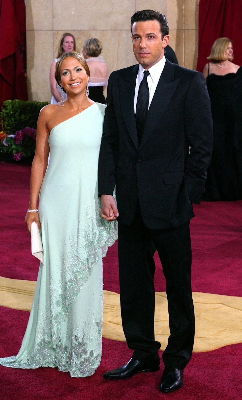 A celebrity couple poses on the red carpet. The woman wears a one-shoulder gown, and the man is in a classic tuxedo