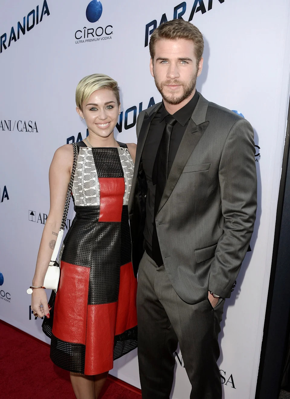 Two people pose on a red carpet. One wears a checkered dress, the other a gray suit