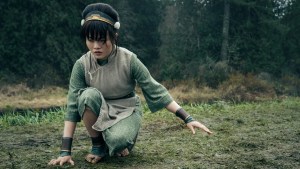 Miya Cech as Toph in "Avatar: The Last Airbender" (dressed in Toph's green clothing and gold/green headband from the animated series) crouches with one hand to the grass and the other hand held out in a wooded clearing