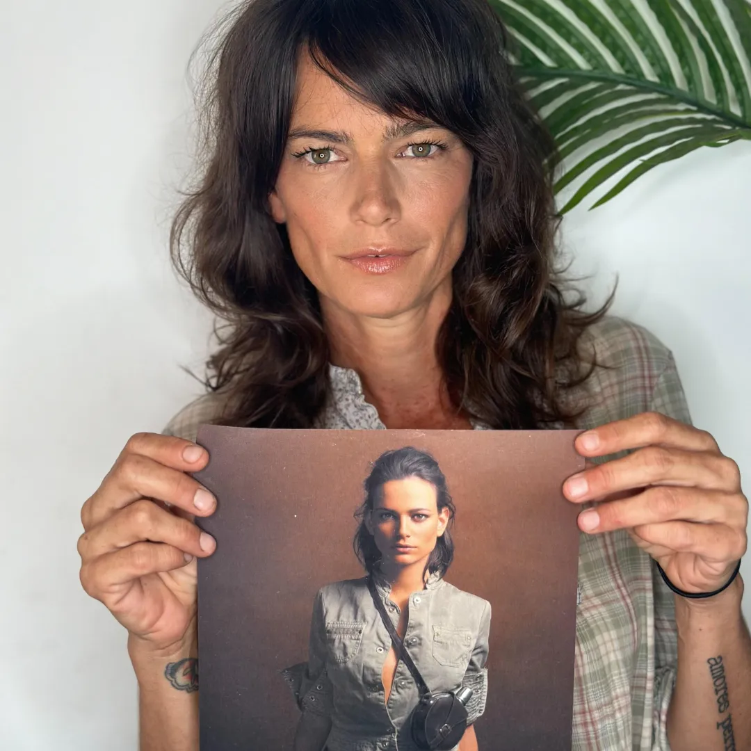 A woman with wavy brown hair holds a photo of herself as a younger woman against a plain background, with part of a green plant visible behind her.
