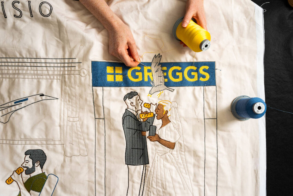 Somebody stitching part of a huge tapestry featuring pictures of people eating Greggs sausage rolls