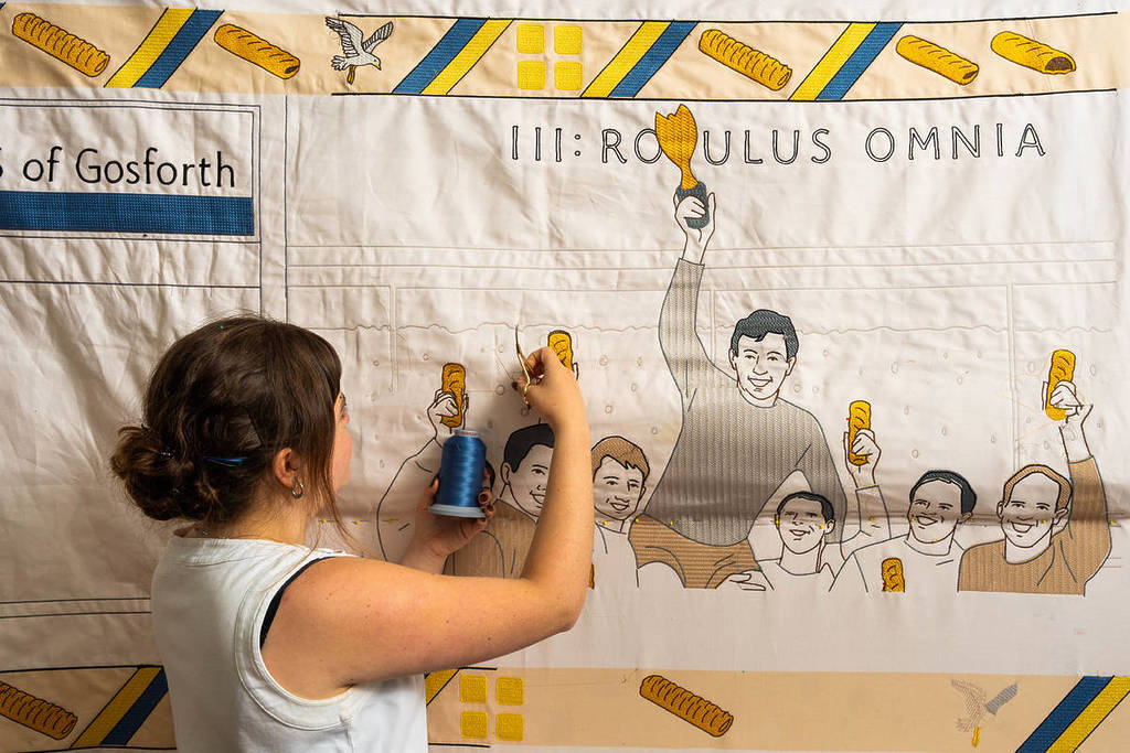 Somebody stitching part of a huge tapestry featuring pictures of people eating Greggs sausage rolls