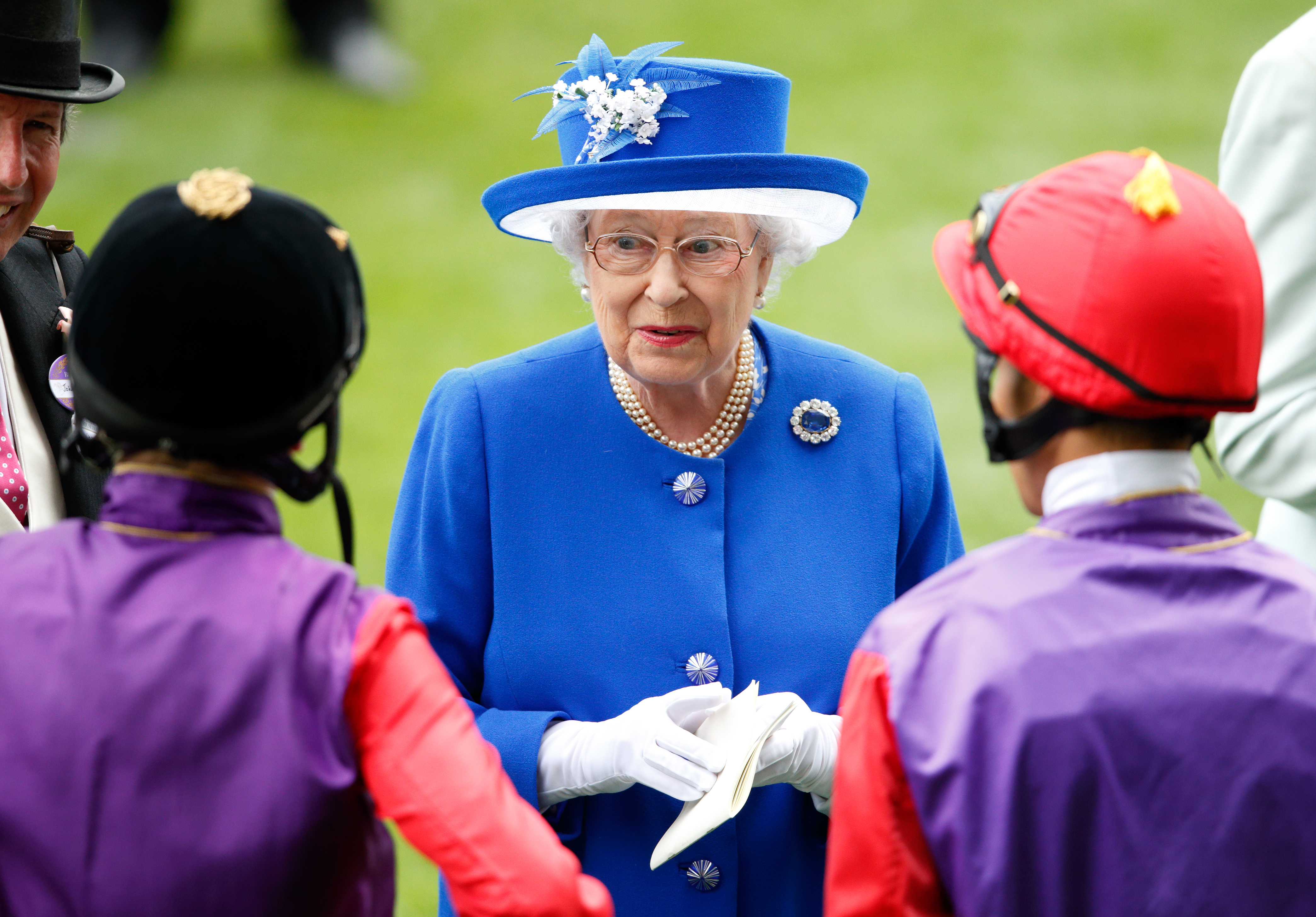 Queen Elizabeth wearing a blue coat and hat talking to jockeys at Royal Ascot