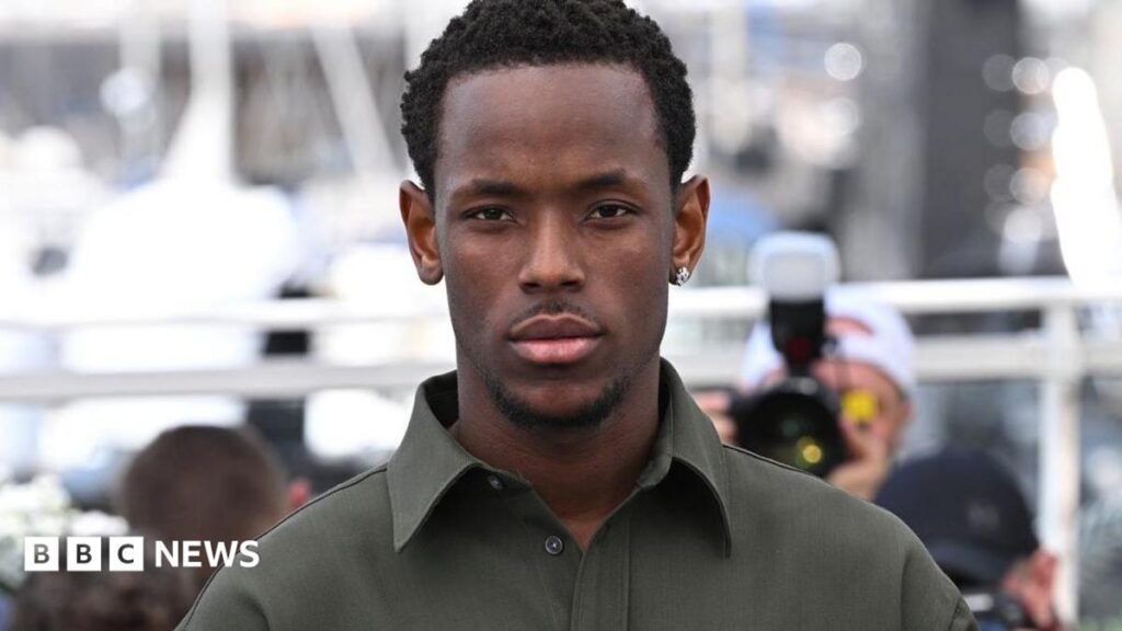 Micheal Ward, surrounded by photographers and wearing a dark green shirt and earring, looks straight into the camera