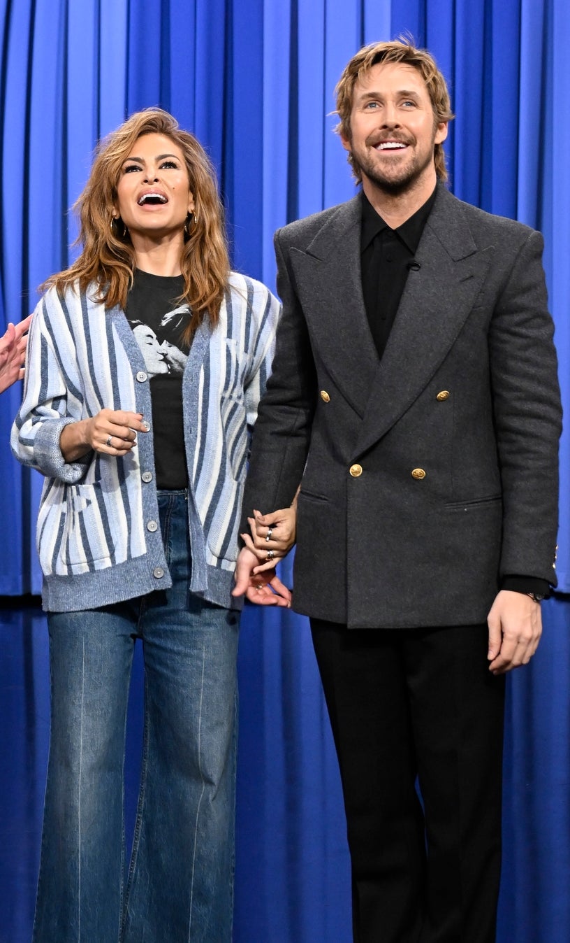 Three people stand on the set of a talk show, with one wearing a suit, another in a striped jacket and flared jeans, and the third in a sweater and pants