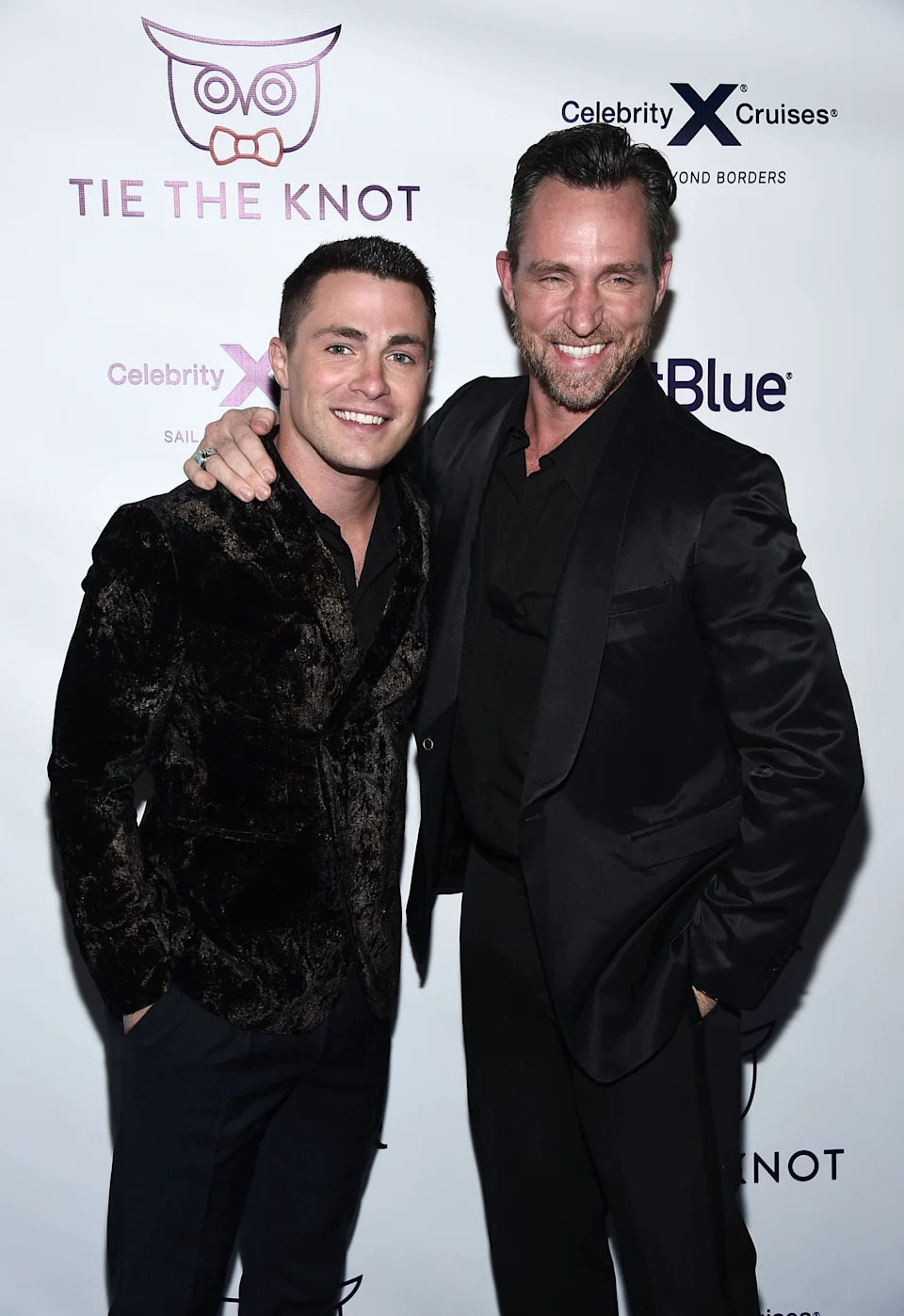 Two people smiling and posing together on a "Tie The Knot" event red carpet, both wearing stylish dark, formal outfits