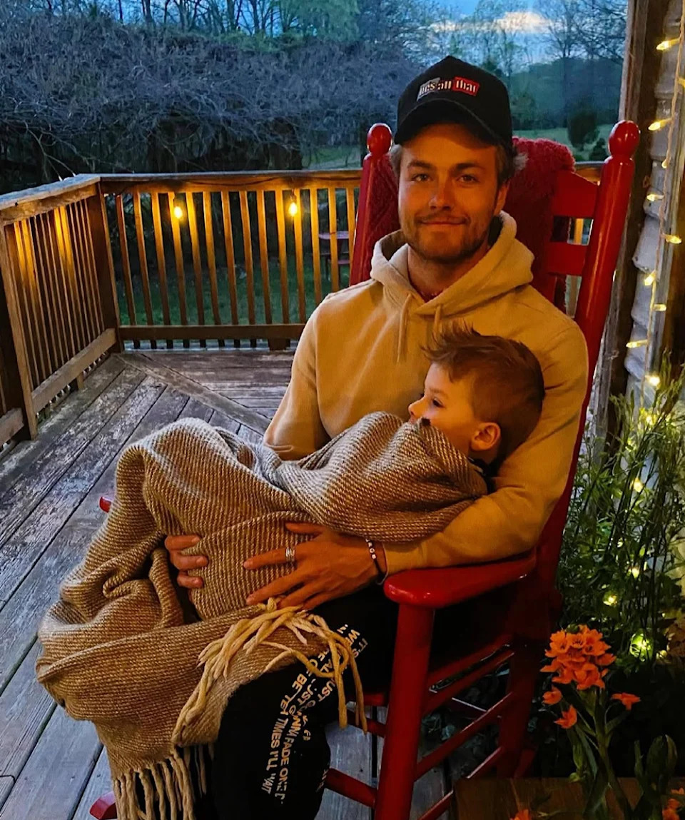 A person wearing a hoodie and cap sits on a porch in a rocking chair, holding a child wrapped in a blanket. They appear content and relaxed