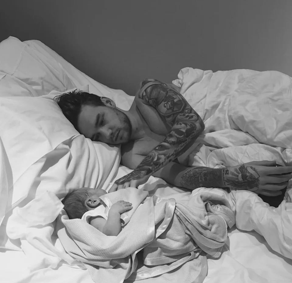 A person with tattoos lies in bed, gently touching a sleeping baby