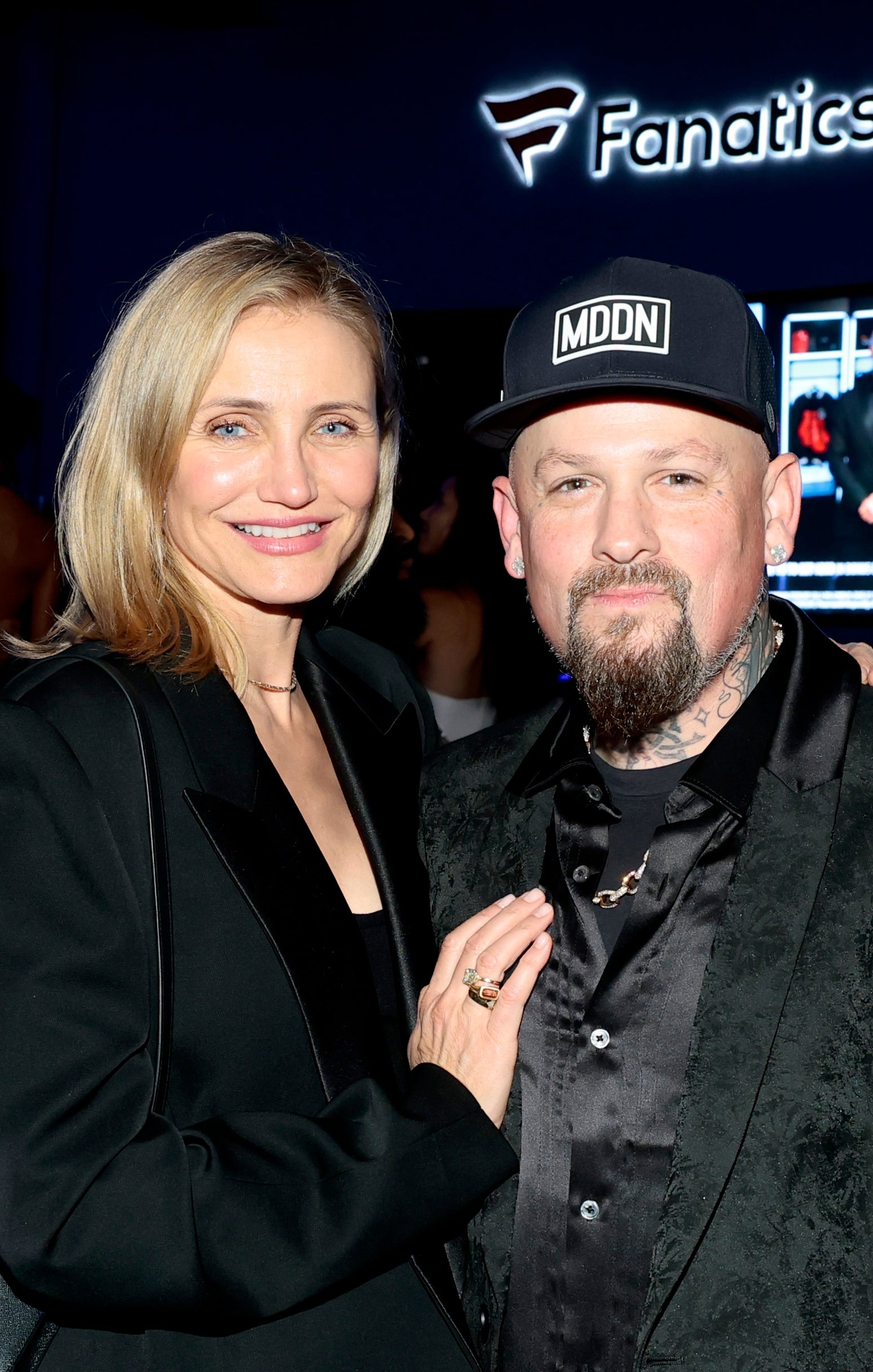A couple poses together at an event, the woman in an elegant blazer and the man in a patterned jacket and cap, with signage in the background