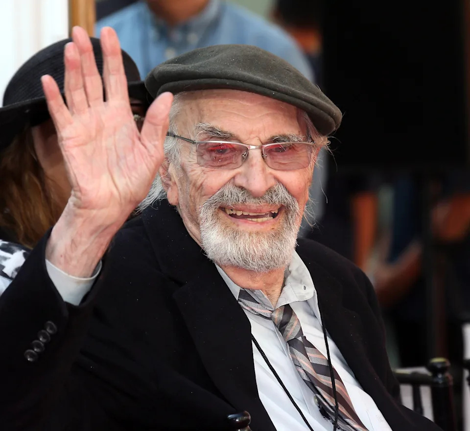Elderly man with beard and glasses, wearing a flat cap and suit, waves and smiles at an outdoor event