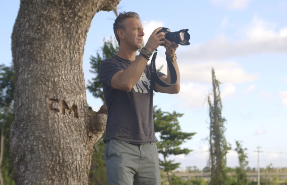 A person stands outdoors, focusing on a camera, with trees and a clear sky in the background