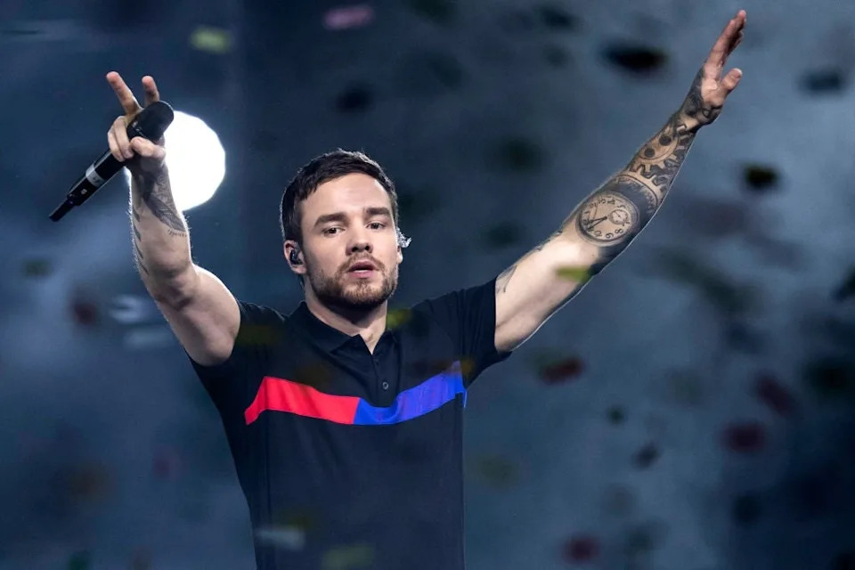 Person performing on stage, holding a microphone, arms raised, with visible tattoos on one arm, wearing a polo shirt with red and blue stripes