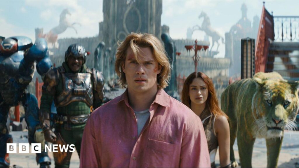 Masters of the Universe still. A group of superhero characters and a CGI lion are stood in a line in front of a tall building.