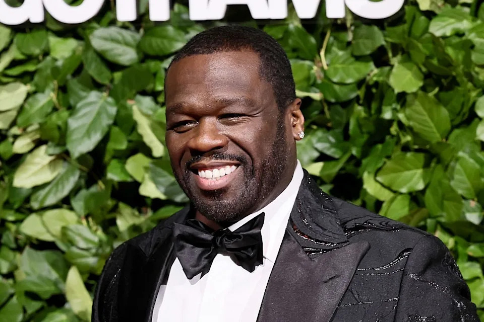 Man smiling in front of leafy backdrop wearing a black tuxedo with a bow tie
