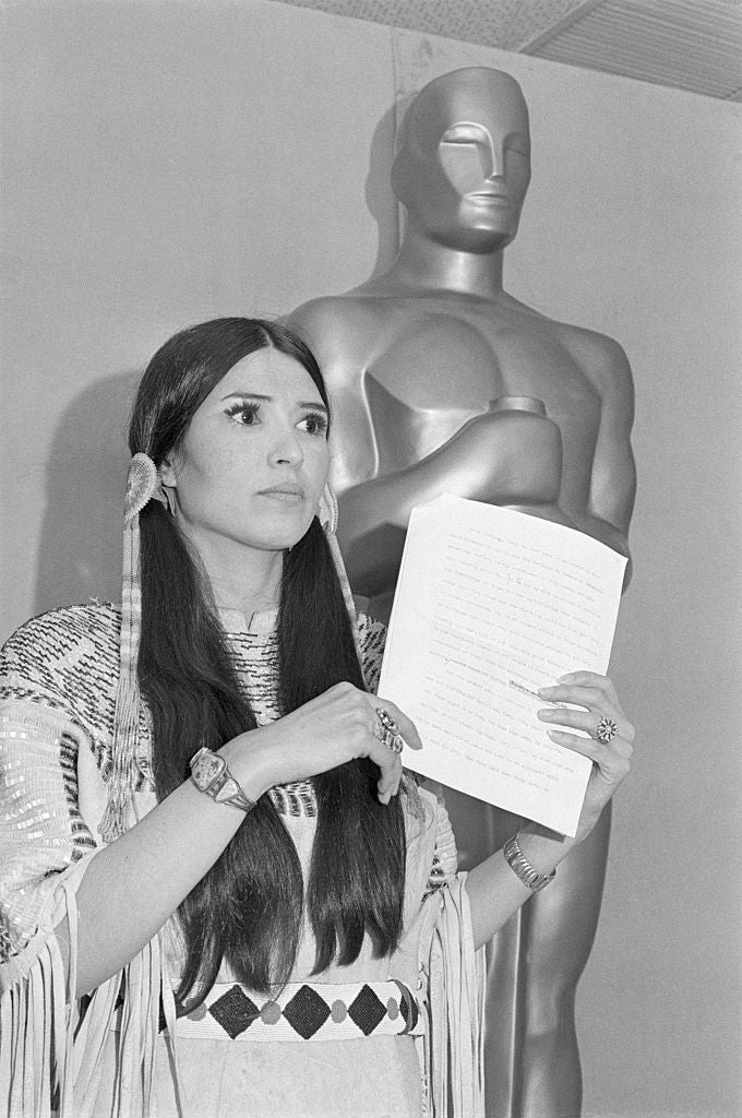 Sacheen Littlefeather  in traditional attire holding a speech in front of a large statue at an awards event