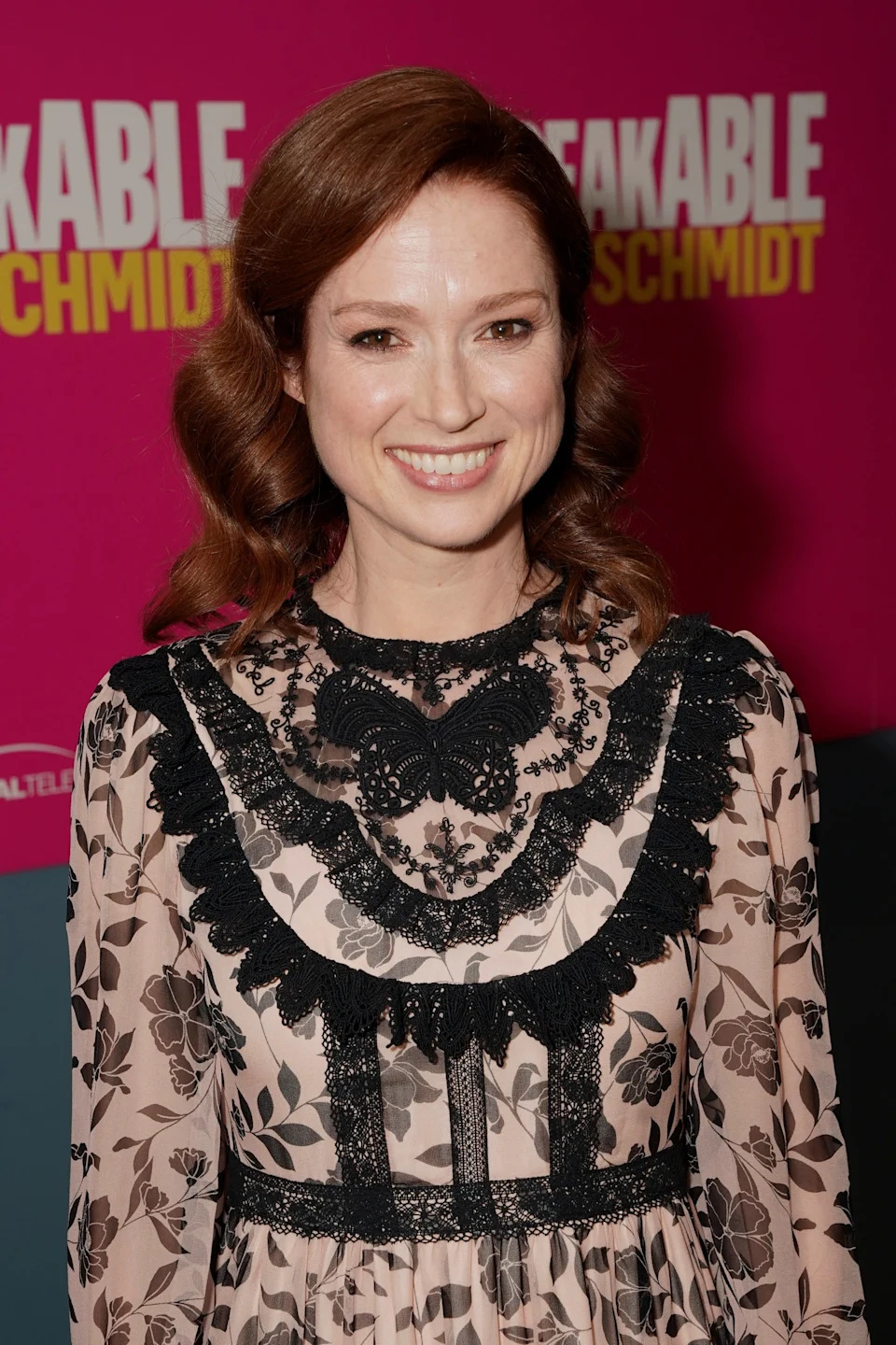 Ellie Kemper smiling at a red carpet event with an "Unbreakable Kimmy Schmidt" backdrop