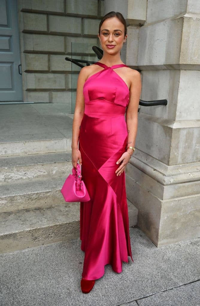 Lady Amelia Windsor in a pink satin gown and red heels stands outside a building, holding a pink purse.