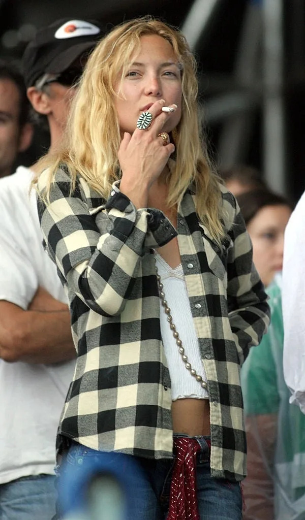 Person with long hair wearing a checkered shirt and holding a cigarette, standing outdoors at an event