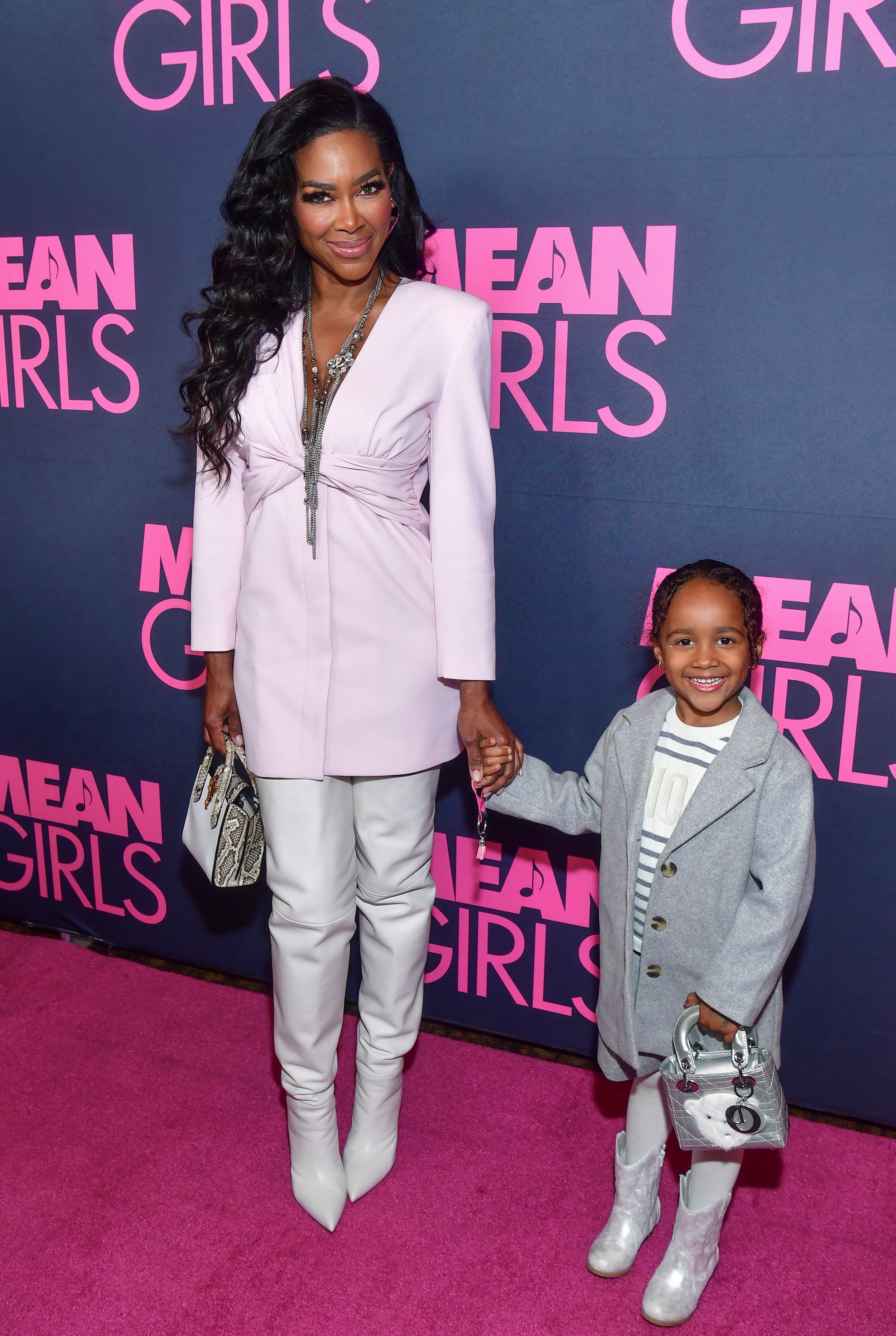 Woman and child on red carpet, woman in tailored dress and high boots, child in coat and striped dress holding a bag