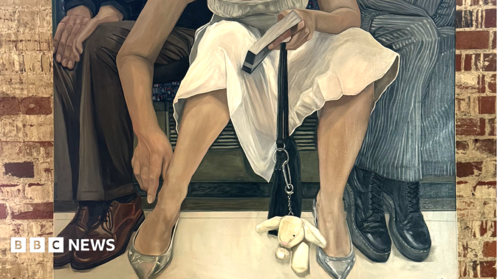 A woman stands next to a large piece of art. She is smiling and has dark hair that has been tied back. She is wearing a black blouse and a white skirt. The painting shows a woman sat on a tube who has spread her legs wide. She is encroaching on two men wearing suits sat beside her who have angled their legs away from her. The woman is wearing a white summer dress and has a black bag with a rabbit key chain attached to it that rests on the floor between her legs. She holds a book in one hand and her other hand is down toward her ankle.