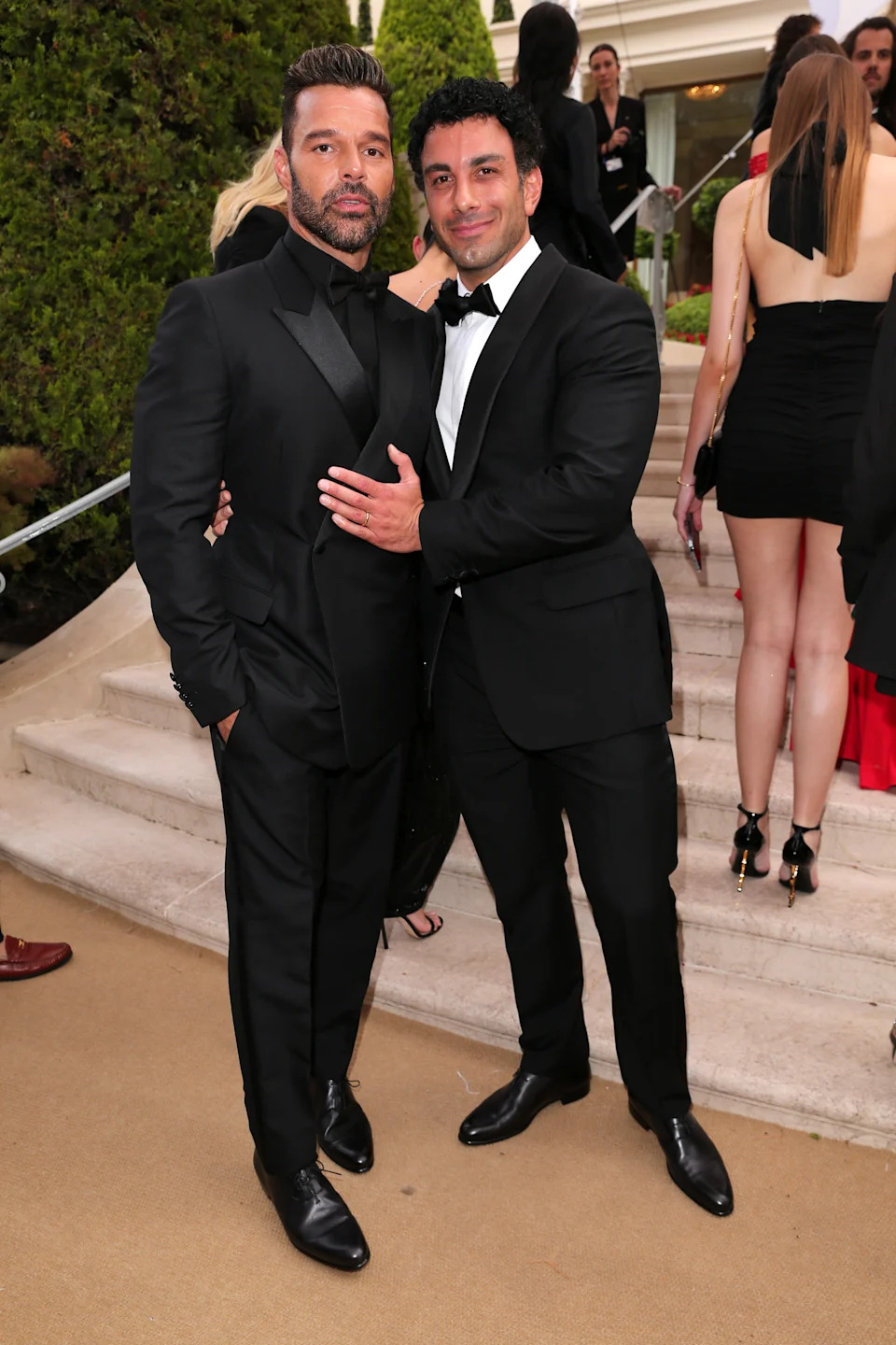 Two people in elegant black tuxedos stand together on a formal event's outdoor steps, surrounded by other attendees