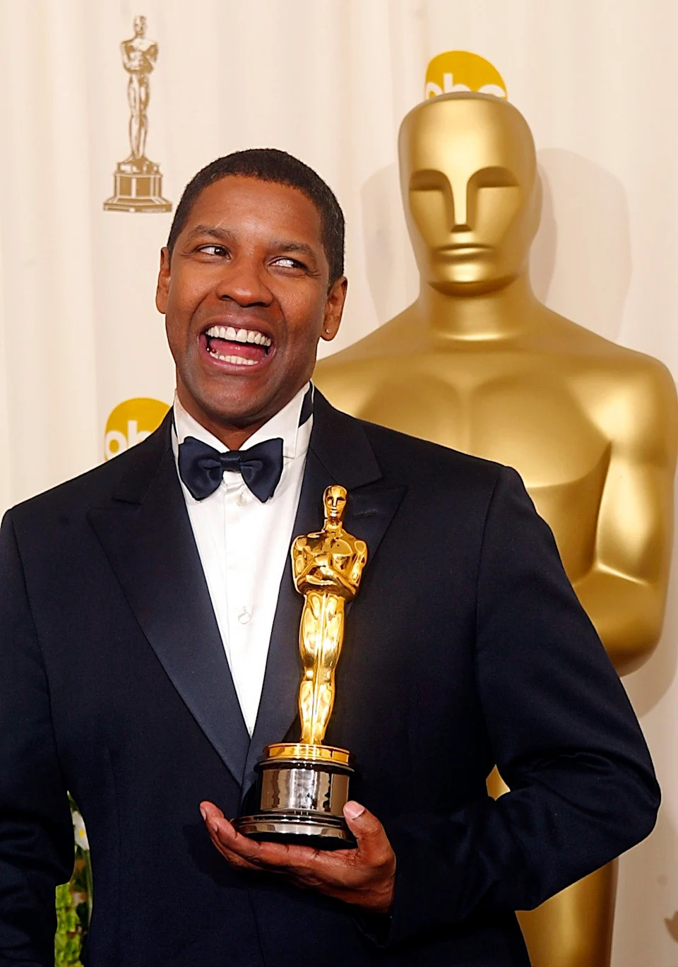 A person smiling broadly, holding an Oscar statue, standing in front of a larger Oscar statue. They are wearing a formal black tuxedo