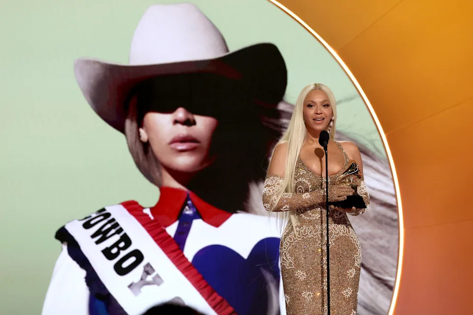 A woman in an elegant off-the-shoulder gown holds an award on stage, with a large image of a woman in a cowboy hat in the background