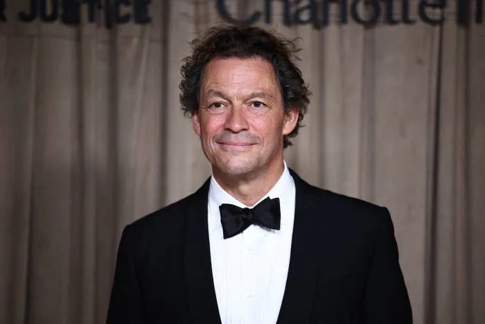 Dominic West in a tuxedo with a bow tie at a formal event. Neutral expression, standing in front of a backdrop with text