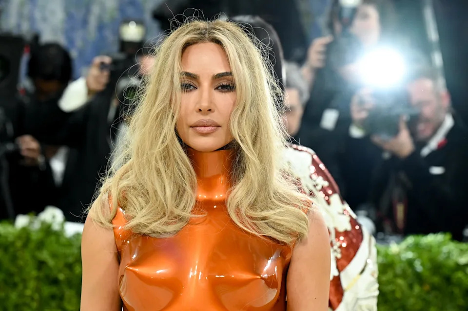 Kim Kardashian on the Met Gala red carpet in shiny, form-fitting attire with flowing hair, surrounded by photographers and flashes