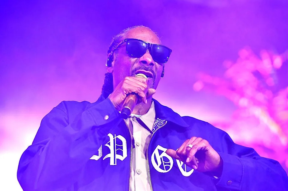 Person in sunglasses and casual jacket performs on stage, holding a microphone