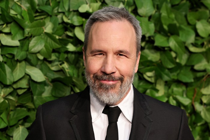 Denis Villeneuve wearing a suit and smiling, in front of a green leafy backdrop.