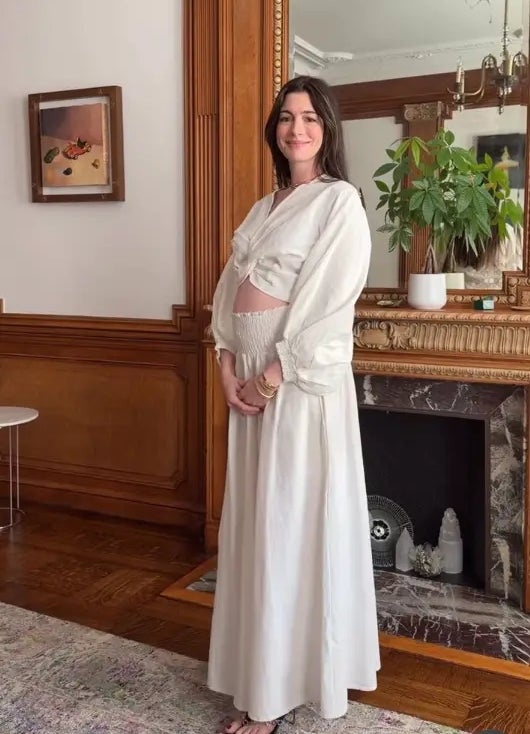 A person stands in an elegant room, wearing a flowing white outfit with long sleeves and a skirt. They are posing in front of a decorative fireplace