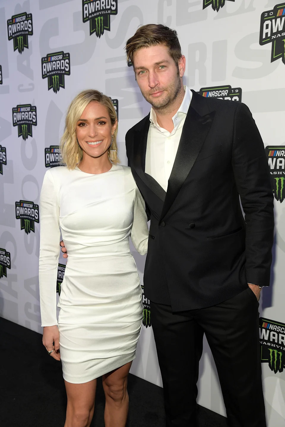 Two people at an event, one wearing a sleek white dress and the other in a sharp black tuxedo, posing on a red carpet