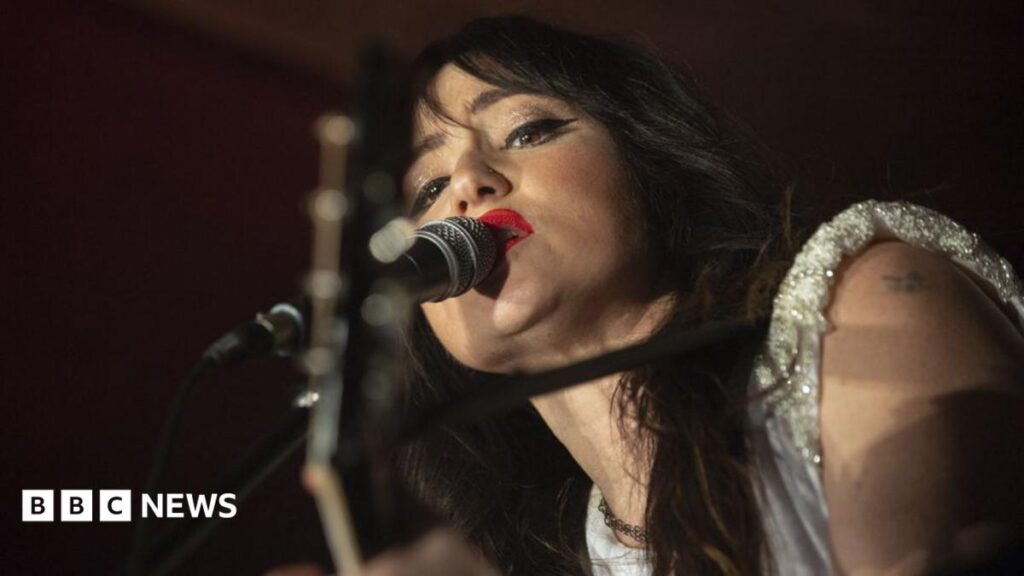 Singer KT Tunstall with long dark hair and a silver top sings into a microphone
