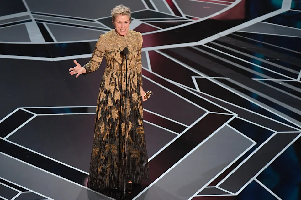 Frances McDormand in patterned long dress on stage, holding an award, speaking passionately at a formal event. Geometric floor pattern featured
