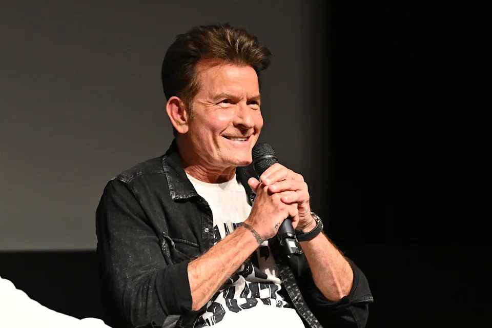 Charlie Sheen smiling and holding a microphone, wearing a casual jacket over a graphic t-shirt, seated on stage during a panel discussion