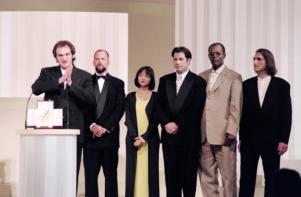 Quentin Tarantino and a Group of six people on stage wearing formal attire, with the man at a podium speaking, possibly at an award ceremony